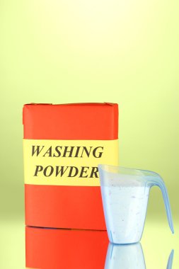 Box of washing powder with blue measuring cup, on green background close-up clipart