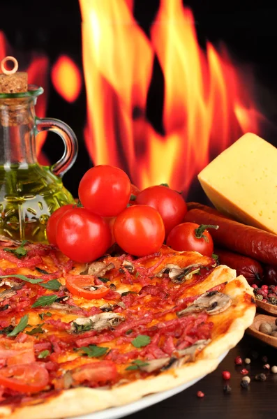 Delicious pizza, salami, tomatoes and spices on wooden table on flame background — Stock Photo, Image