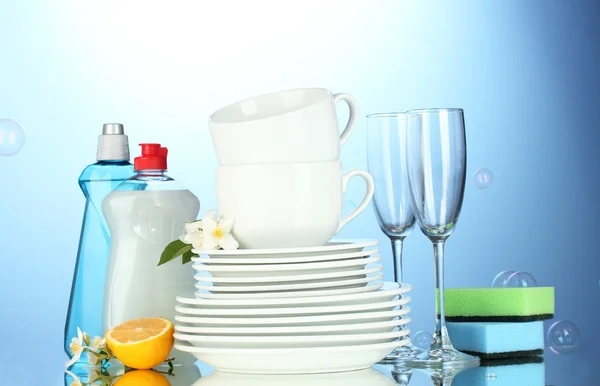 Assiettes propres vides, verres et tasses avec liquide vaisselle, éponges et citron sur fond bleu — Photo