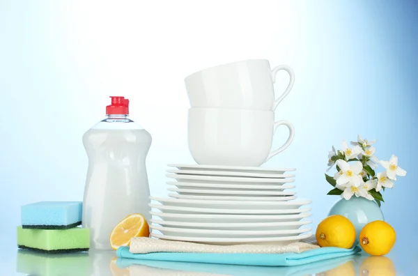 Platos y tazas limpias vacías con líquido lavavajillas, esponjas y limón sobre fondo azul —  Fotos de Stock