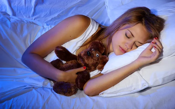 Young beautiful woman with toy bear sleeping on bed in bedroom — Stock Photo, Image