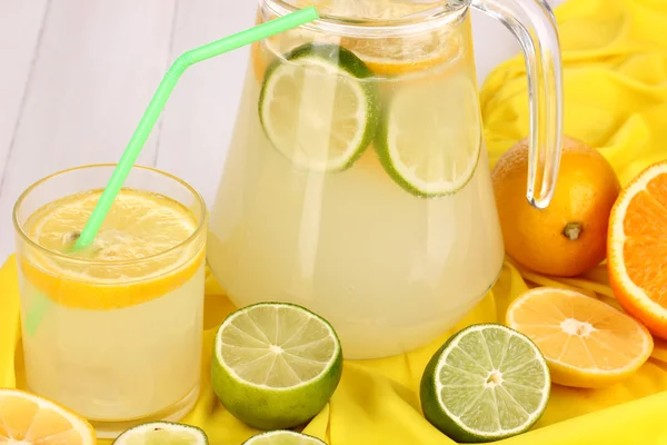 Zitruslimonade im Glas und Krug mit Zitrusfrüchten auf gelbem Stoff auf weißem Holztisch in Großaufnahme — Stockfoto