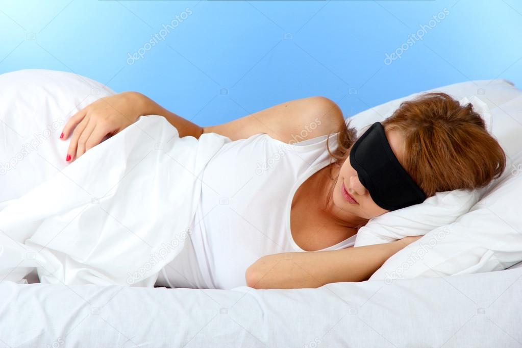 young beautiful woman sleeping in bed with eye mask on blue background