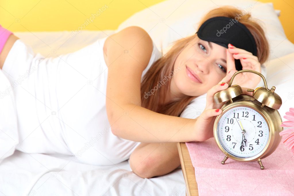young beautiful woman lying on bed with eye mask and alarm clock, on yello