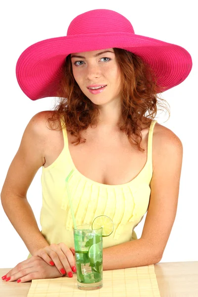 Sorridente bella ragazza seduta a tavola con cappello da spiaggia e cocktail isolato su bianco — Foto Stock