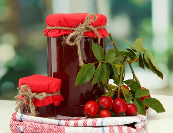 Potten met hip rozen jam en rijpe bessen, op houten tafel — Stockfoto