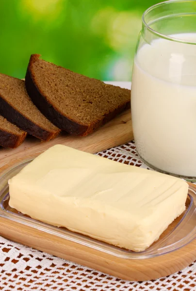 Mantequilla sobre soporte de madera rodeada de pan y leche sobre fondo natural — Foto de Stock