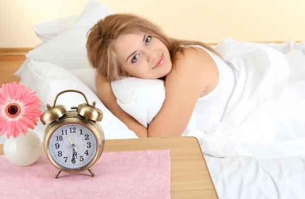 Young beautiful woman lying on bed with alarm clock in bedroom — Stock Photo, Image