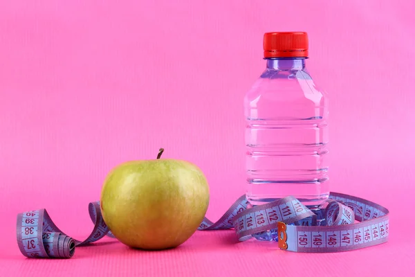 Garrafa de água, maçã e fita métrica em fundo rosa — Fotografia de Stock