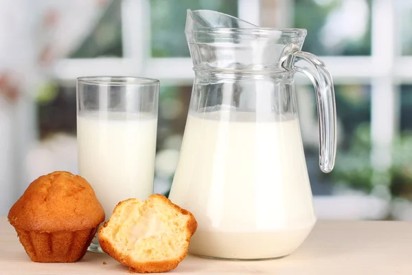 Pitcher and glass of milk with muffins on wooden table on window background — Stock Photo, Image