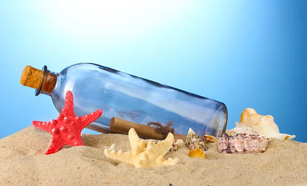 Glass bottle with note inside on sand, on blue background — Stock Photo, Image