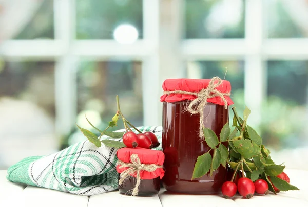 Potten met hip rozen jam en rijpe bessen, op houten tafel — Stockfoto