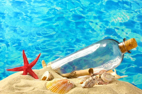 Glass bottle with note inside on sand, on blue sea background — Stock Photo, Image