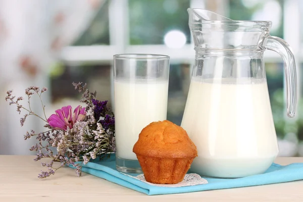 Dzban i szklankę mleka z babeczki na drewnianym stole na tle okna — Zdjęcie stockowe