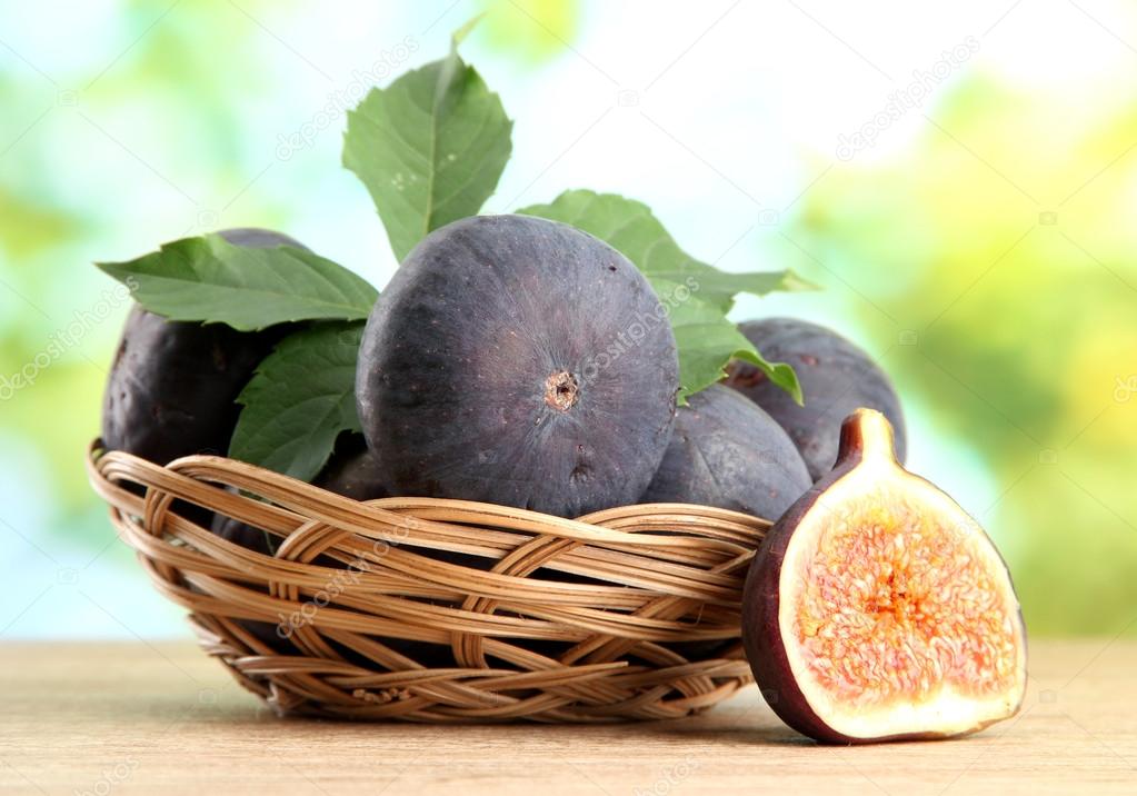 Ripe sweet figs with leaves in basket, on wooden table, on green background