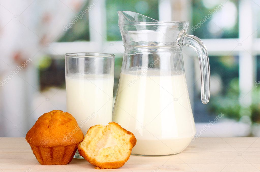 Pitcher and glass of milk with muffins on wooden table on window background