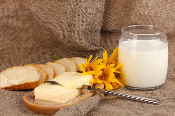 Mantequilla sobre soporte de madera rodeada de pan y leche sobre fondo de saco — Foto de Stock