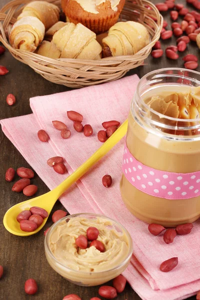 Delicious peanut butter in jar with baking on napkin on wooden table close- — Stock Photo, Image