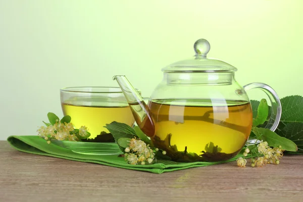 Tetera y taza con té de tilo y flores en mesa de madera sobre fondo verde —  Fotos de Stock