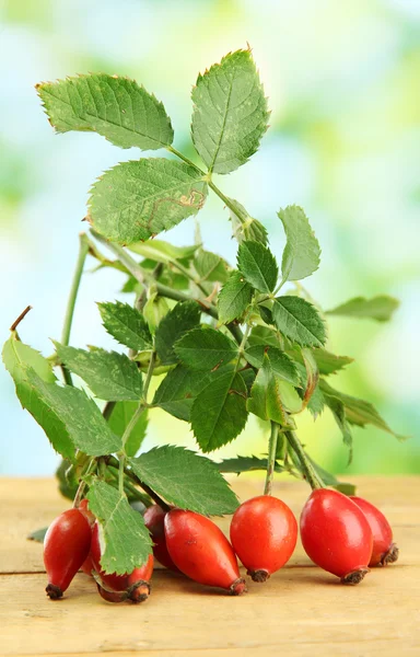 Reife Hüftrosen auf Zweig mit Blättern, auf Holztisch auf grünem Hintergrund — Stockfoto