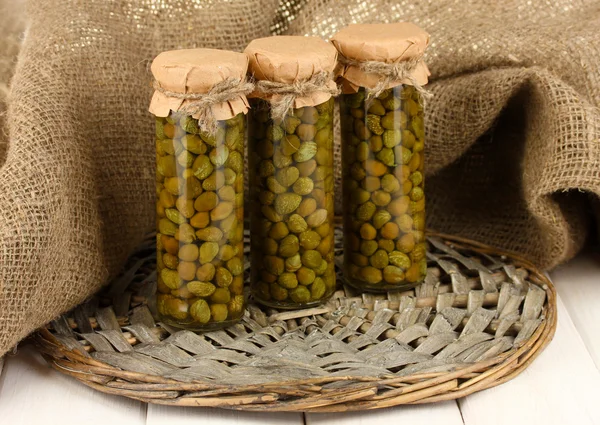 Glass jars with tinned capers in sack on white wooden background — Stock Photo, Image