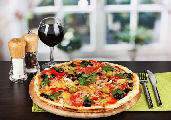 Leckere Pizza mit einem Glas Rotwein und Gewürzen auf einem Holztisch am Fenster — Stockfoto