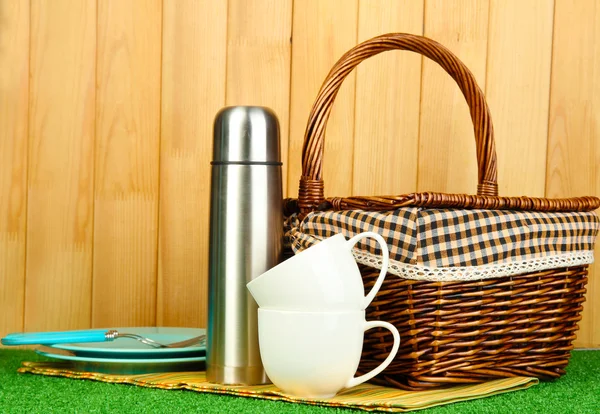 Metallthermoskanne mit Tassen, Tellern und Korb auf Gras auf Holzgrund — Stockfoto