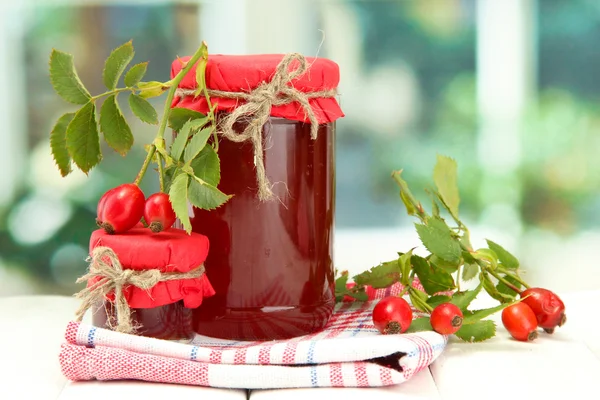 Jars with hip roses jam and ripe berries, on wooden table — Stock Photo, Image