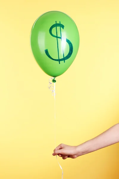 Ręka trzyma zielonego balonika jeden z pokazując Dolar na żółtym tle — Zdjęcie stockowe