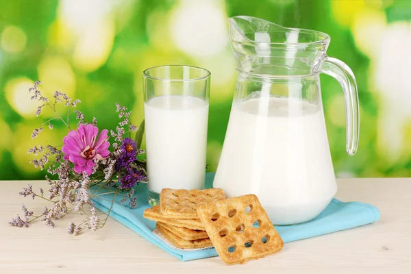 Jarra y vaso de leche con galletas sobre mesa de madera sobre fondo natural — Foto de Stock