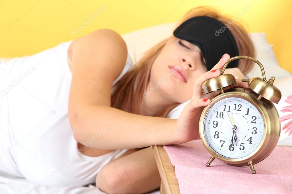 Young beautiful woman lying on bed with eye mask and alarm clock, on yellow background