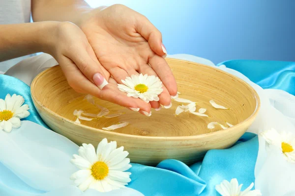 Vrouw handen met houten kom met water met bloemen, op blauwe achtergrond — Stockfoto