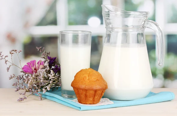 Jarro e copo de leite com muffins na mesa de madeira no fundo da janela — Fotografia de Stock
