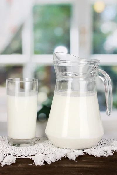 Pitcher and glass of milk on wooden table on window background — Stock Photo, Image