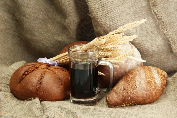 Tankard of kvass and rye breads with ears, on burlap background — Stock Photo, Image