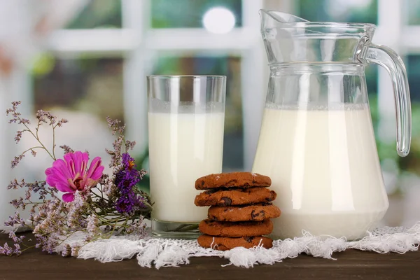 Jarro e copo de leite com biscoitos na mesa de madeira no fundo da janela — Fotografia de Stock
