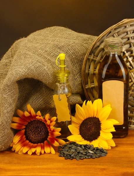 Oil in bottles, sunflowers and seeds, on wooden table on brown background — Stock Photo, Image