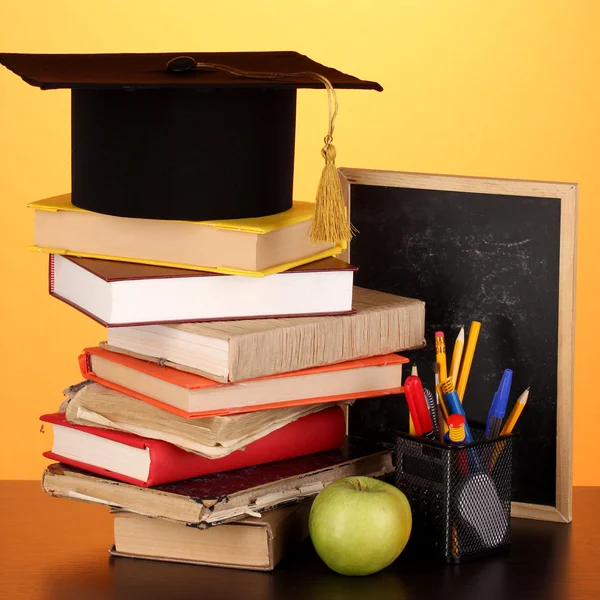 Livros e magister cap contra placa da escola em mesa de madeira em amarelo backg — Fotografia de Stock