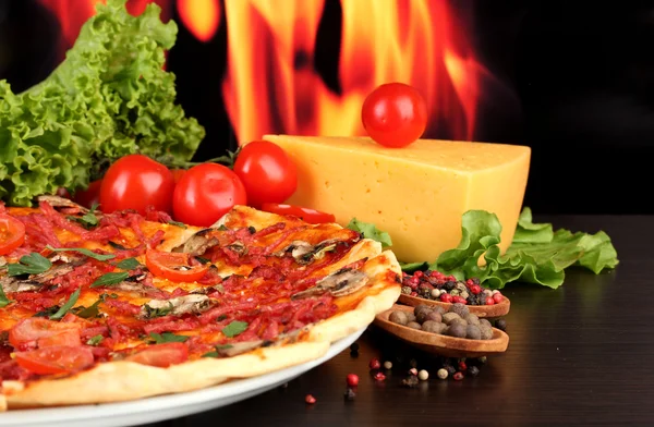 Delicious pizza, salami, tomatoes and spices on wooden table on flame backg — Stock Photo, Image