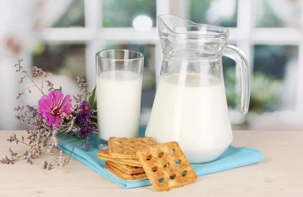 Dzban i szklankę mleka z plików cookie na drewnianym stole na tle okna — Zdjęcie stockowe