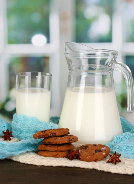 Кувшин и стакан молока с печеньем на крючках трикотаж на деревянном столе на фоне окна — стоковое фото