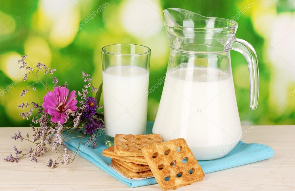Pitcher and glass of milk with cookies on wooden table on natural background