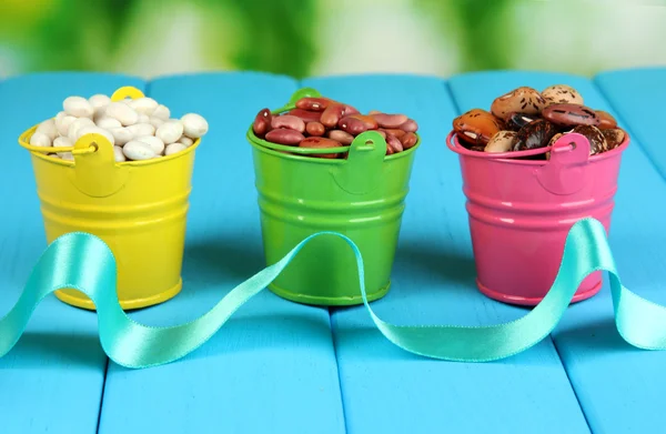 Different types of beans in colored buckets on blue wooden table on natural background — Stock Photo, Image