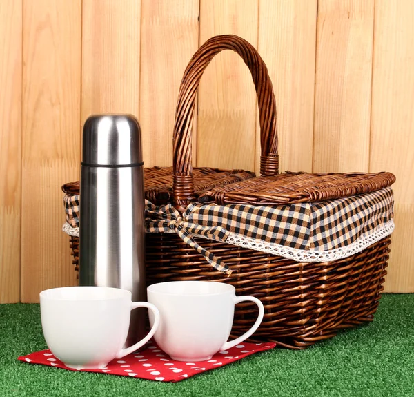 Metal thermos with cups and basket on grass on wooden background — Stock Photo, Image
