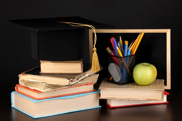 Livros e magister cap contra placa da escola em mesa de madeira sobre fundo preto — Fotografia de Stock