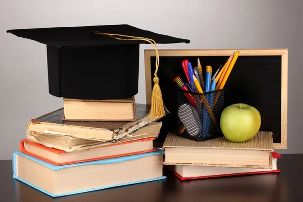 Books and magister cap against school board on wooden table on grey background — Stock Photo, Image