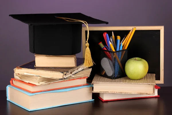 Livros e magister cap contra placa da escola em mesa de madeira no fundo roxo — Fotografia de Stock