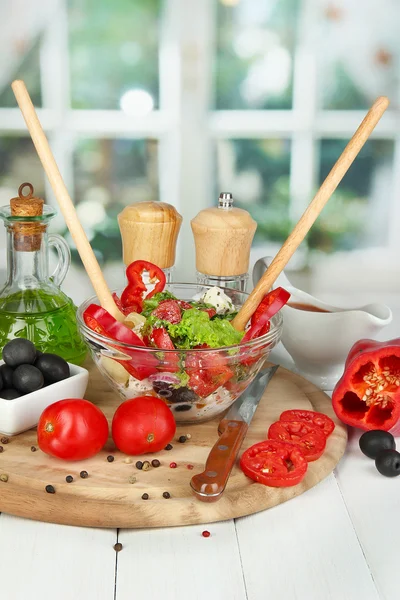 Insalata greca fresca in una ciotola di vetro circondata da ingredienti per cucinare su un tavolo di legno su fondo finestra — Foto Stock