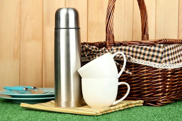 Metal thermos with cups, plates and basket on grass on wooden background — Stock Photo, Image