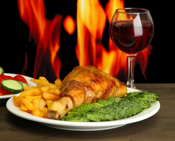 Roast chicken with french fries and cucumbers, glass of wine on wooden table on fire background — Stock Photo, Image